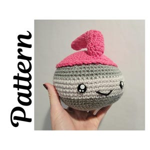 May include: Crocheted curling stone with a smiling face, grey and white striped body, and a pink top and handle. The word "Pattern" is printed vertically in black on the left side. The stone is held in a hand.