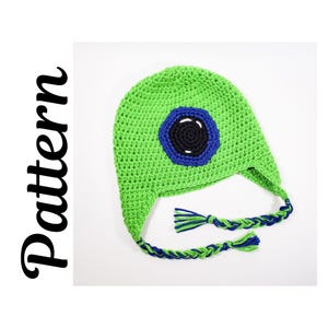 May include: A green crocheted hat with a large eye design in blue and black. The hat has ear flaps and braided ties with green and blue tassels. The word "Pattern" is written vertically in black cursive on the left side.
