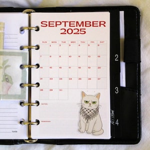 May include: A planner open to the September 2025 calendar page. The calendar is white with red text and a cartoon cat sticker. The planner is black with gold rings and a black elastic band.