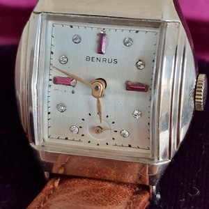 May include: A vintage Benrus wristwatch with a square silver-tone case and a brown leather strap. The white dial features gold-tone hands, a small sub-dial, and hour markers consisting of alternating round clear stones and rectangular red stones.