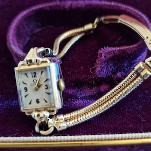 May include: Vintage gold-toned Omega wristwatch with a rectangular face and a white dial. The watch features gold hands and hour markers, and a gold bracelet. The watch is displayed on a purple velvet surface.