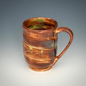 May include: A ceramic mug with a glossy finish featuring a warm, variegated brown glaze with subtle hints of green. The mug has a cylindrical body with horizontal ridges and a curved handle attached to the side, showcasing a hand-thrown appearance.