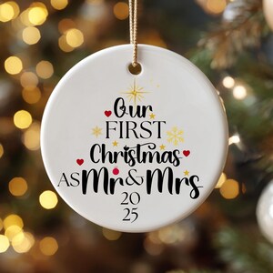 May include: White ceramic Christmas ornament with gold string, featuring the text "Our First Christmas as Mr & Mrs 2025" in black script with red heart accents. The ornament hangs in front of a blurred Christmas tree.