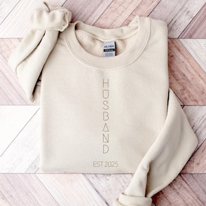 May include: A cream-colored sweatshirt with the word "HUSBAND" printed vertically in a brown font. Below the word is "EST 2025". The sweatshirt is folded on a wooden surface.