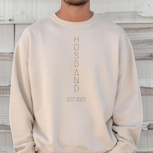 May include: A beige crewneck sweatshirt with the word "HUSBAND" printed vertically in a minimalist font. Below the word is "EST 2025" in a similar font. The sweatshirt is a solid color and has long sleeves.