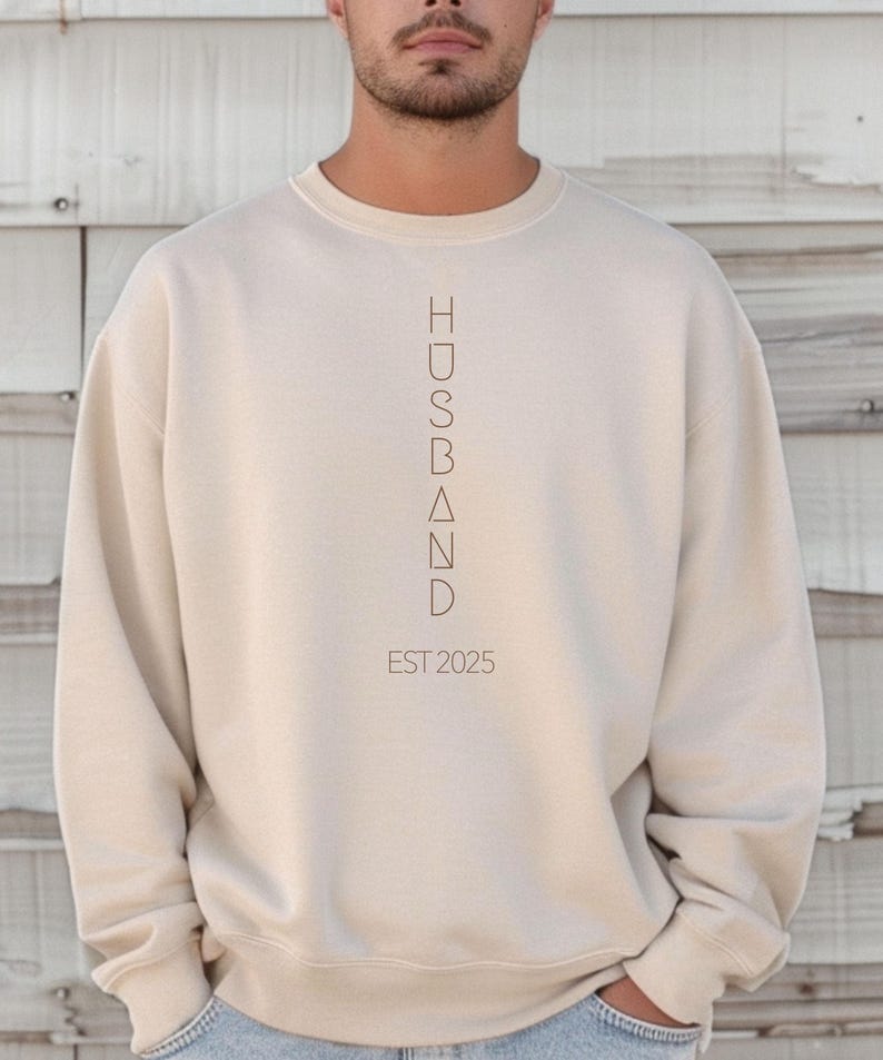 May include: A beige crewneck sweatshirt with the word "HUSBAND" printed vertically in a minimalist font. Below the word is "EST 2025" in a similar font. The sweatshirt is a solid color and has long sleeves.