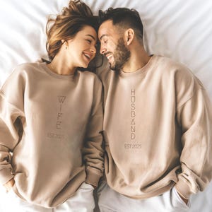 May include: Two people wearing matching beige sweatshirts. One sweatshirt says "WIFE EST 2025", the other says "HUSBAND EST 2025". The couple is smiling and lying on a white surface.
