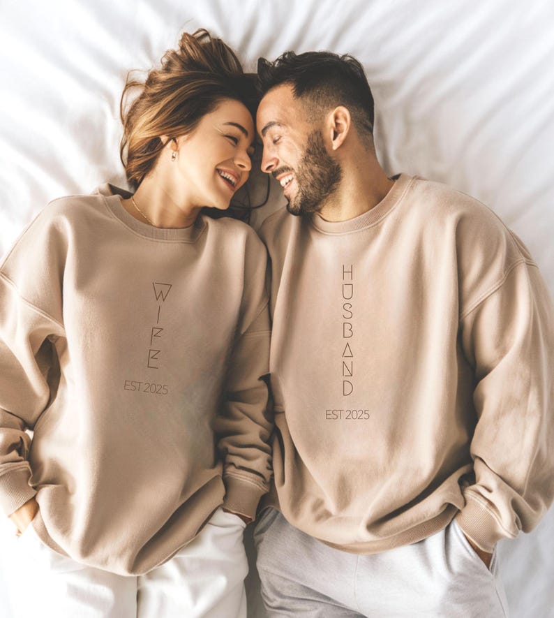 May include: Two people wearing matching beige sweatshirts. One sweatshirt says "WIFE EST 2025", the other says "HUSBAND EST 2025". The couple is smiling and lying on a white surface.