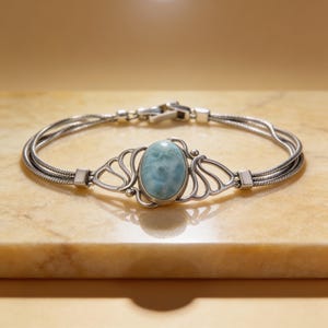 May include: A silver bracelet featuring a central oval light blue stone set in a decorative metal frame. The band consists of three thin, flexible silver chains that connect to the central setting, secured by a silver clasp at the back.
