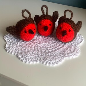 May include: Three handmade crochet robin ornaments. Each bird has a red breast, brown head and wings, and a loop for hanging. They are displayed on a white, crocheted doily.