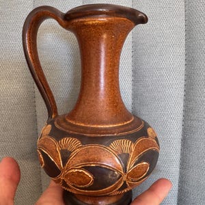 Op de afbeelding: Een bruine keramische kan met een gebogen handvat en een tuit. De kan heeft een donkerbruine basis en een lichtbruine body met een decoratief bloemmotief rond de basis. De kan wordt in de hand van een persoon gehouden.