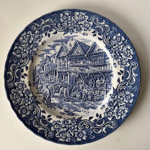 Puede incluir: Un plato decorativo azul y blanco con una ilustración detallada de una escena histórica de una ciudad. El plato presenta un borde floral y un borde festoneado. La imagen central representa edificios, personas y un carro tirado por caballos.