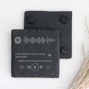 May include: Two square black slate coasters. One coaster features a Spotify code, song title, and artist name: "I don't want to miss a thing" by Aerosmith. The other coaster has two black circular pads on the back.
