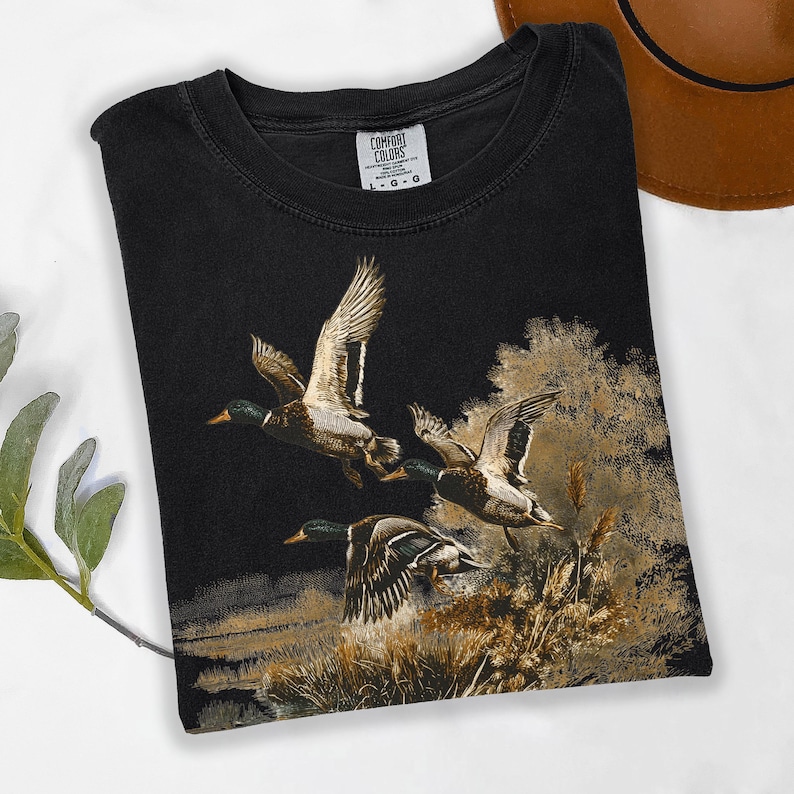 May include: Black t-shirt with a detailed illustration of three ducks in flight over a marsh. The ducks are depicted with brown, white, and green feathers. The shirt has a crew neck and the Comfort Colors logo.