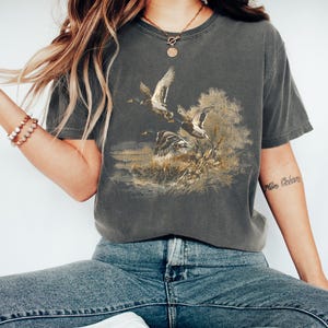 May include: Dark grey t-shirt with a detailed illustration of ducks in flight over a marshy landscape. The artwork is in brown, gold, and green, giving a vintage look. The shirt has a classic crew neck and short sleeves.