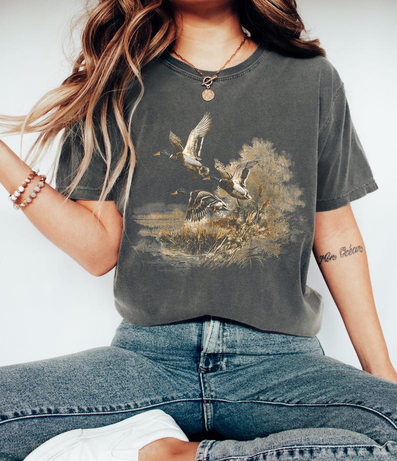 May include: Dark grey t-shirt with a detailed illustration of ducks in flight over a marshy landscape. The artwork is in brown, gold, and green, giving a vintage look. The shirt has a classic crew neck and short sleeves.