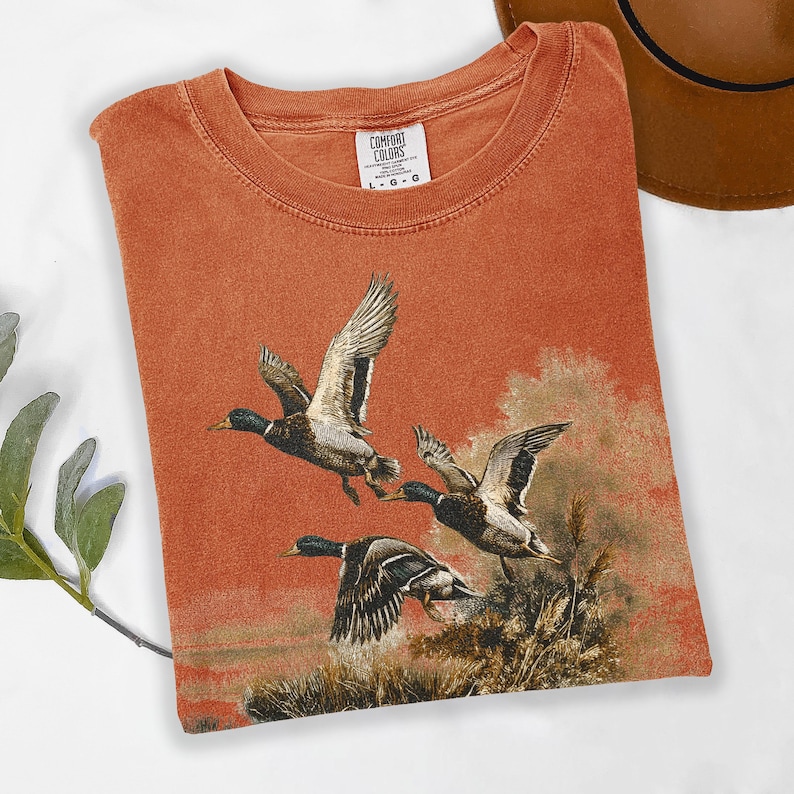 May include: Burnt orange t-shirt with a detailed illustration of three ducks in flight. The ducks are depicted with brown, black, and white feathers, set against a backdrop of reeds and a muted landscape. The shirt has a crew neck and the tag reads "COMFORT COLORS".