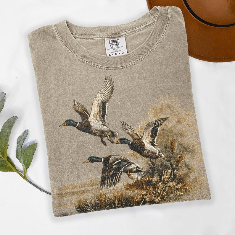 May include: A beige Comfort Colors t-shirt with a detailed illustration of three ducks in flight. The ducks have brown, black, and white feathers. The shirt has a crew neck and the Comfort Colors logo.
