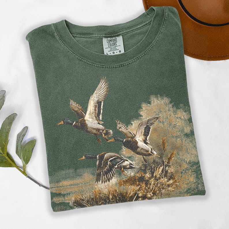 May include: Green Comfort Colors t-shirt with a detailed illustration of three ducks in flight over a marsh. The ducks are depicted with brown and white feathers, set against a backdrop of reeds and water. The shirt has a crew neck.