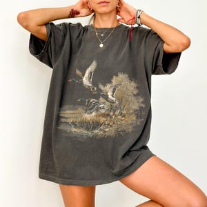 May include: Dark gray oversized t-shirt featuring a nature scene with flying ducks over a body of water. The graphic print includes trees and reeds in shades of brown and tan. The shirt has short sleeves and a relaxed fit.