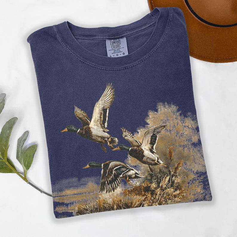 May include: A navy blue Comfort Colors t-shirt with a print of three ducks flying. The ducks are brown and white, set against a backdrop of reeds and a dark blue sky. The shirt has a crew neck and the Comfort Colors label is visible.