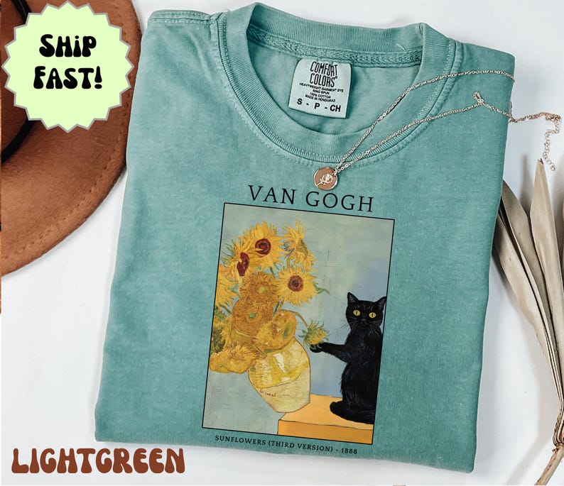 May include: Light green t-shirt featuring a print of Van Gogh's "Sunflowers" with a black cat. The shirt has a crew neck and short sleeves. A silver necklace with a small pendant is on the shirt. The text "VAN GOGH" is above the image.