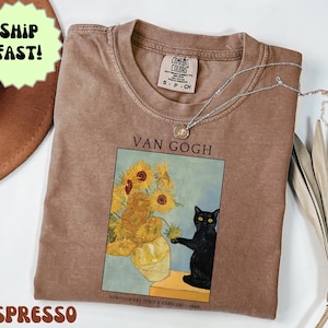 May include: Espresso-colored t-shirt featuring a print of Van Gogh's "Sunflowers" with a black cat. The shirt has a crew neck and a small tag. A silver necklace with a pendant is draped across the shirt.
