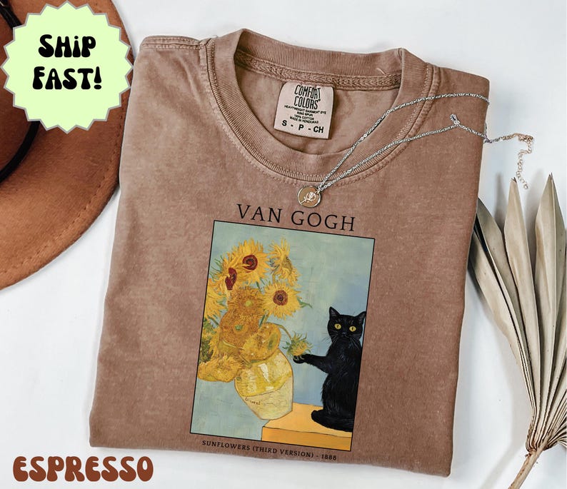 May include: Espresso-colored t-shirt featuring a print of Van Gogh's "Sunflowers" with a black cat. The shirt has a crew neck and a small tag. A silver necklace with a pendant is draped across the shirt.
