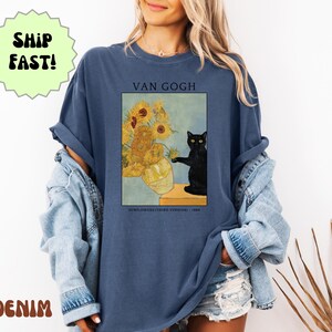 May include: A denim blue t-shirt featuring a print of Van Gogh's "Sunflowers" with a black cat. The text "VAN GOGH" is above the image, and "SUNFLOWERS (THIRD VERSION) - 1888" is below. The shirt has rolled-up sleeves.