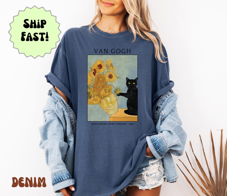 May include: A denim blue t-shirt featuring a print of Van Gogh's "Sunflowers" with a black cat. The text "VAN GOGH" is above the image, and "SUNFLOWERS (THIRD VERSION) - 1888" is below. The shirt has rolled-up sleeves.