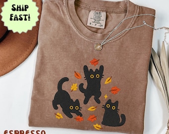 Puede incluir: Camiseta color espresso con tres ilustraciones de gatos negros con ojos amarillos y hojas de otoño. La camiseta tiene cuello redondo y mangas cortas. Un collar plateado con un colgante está sobre la camiseta.