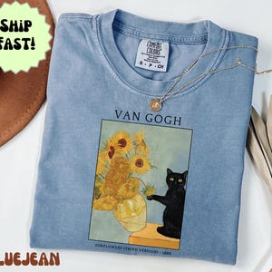 May include: A light blue Comfort Colors t-shirt featuring a print of Van Gogh's "Sunflowers" painting with a black cat. The shirt has a crew neck and short sleeves. A gold necklace is draped across the shirt.