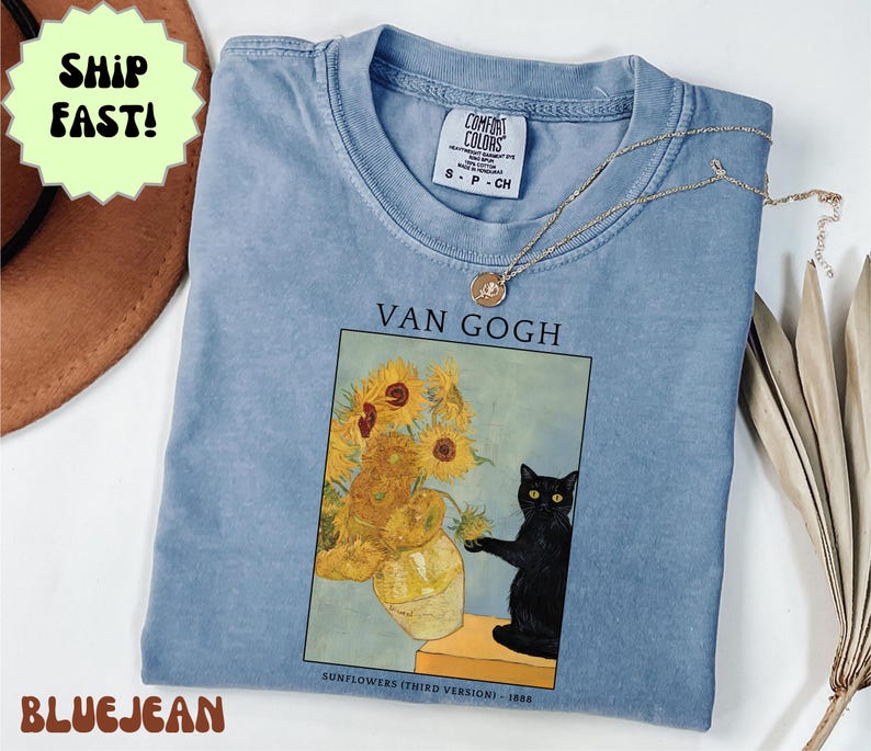 May include: A light blue Comfort Colors t-shirt featuring a print of Van Gogh's "Sunflowers" painting with a black cat. The shirt has a crew neck and short sleeves. A gold necklace is draped across the shirt.