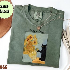 May include: Moss green t-shirt featuring a print of Van Gogh's "Sunflowers" with a black cat. The text "VAN GOGH" is above the image, and "SUNFLOWERS (THIRD VERSION) - 1888" is below. A gold necklace is draped across the shirt.