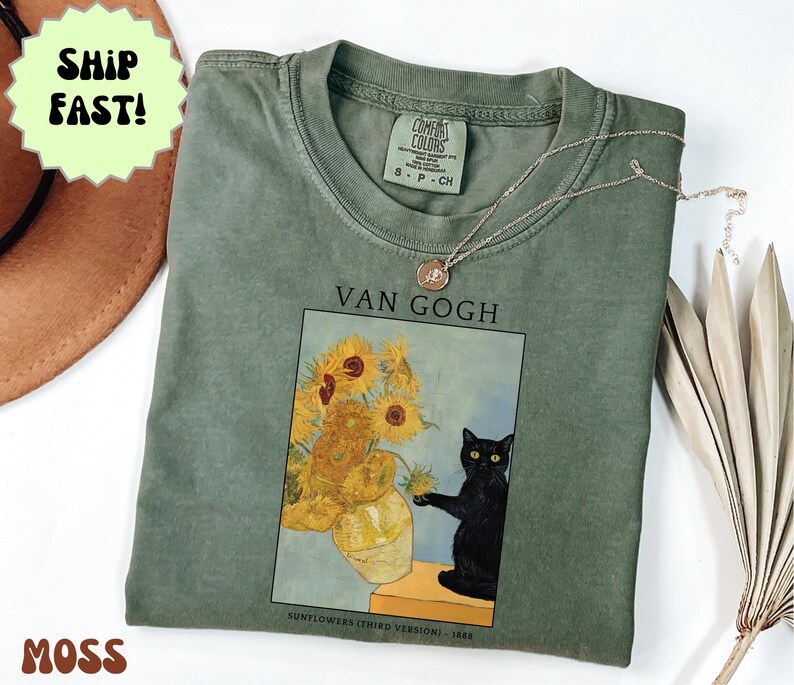 May include: Moss green t-shirt featuring a print of Van Gogh's "Sunflowers" with a black cat. The text "VAN GOGH" is above the image, and "SUNFLOWERS (THIRD VERSION) - 1888" is below. A gold necklace is draped across the shirt.