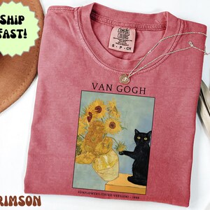 May include: Crimson-colored t-shirt featuring a print of Van Gogh's "Sunflowers" with a black cat. The shirt has a crew neck and short sleeves. A silver necklace with a pendant is draped across the shirt. The text "VAN GOGH" is above the artwork.
