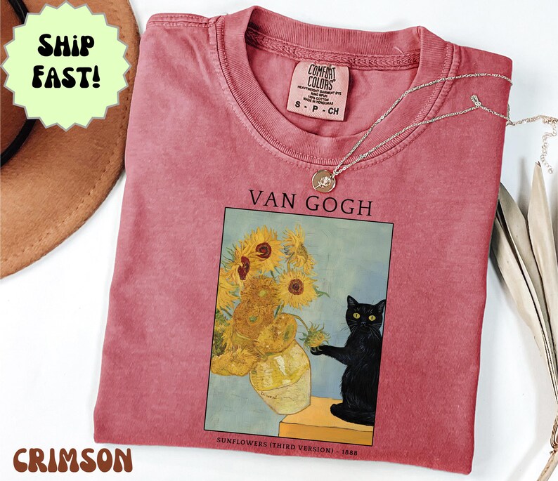 May include: Crimson-colored t-shirt featuring a print of Van Gogh's "Sunflowers" with a black cat. The shirt has a crew neck and short sleeves. A silver necklace with a pendant is draped across the shirt. The text "VAN GOGH" is above the artwork.