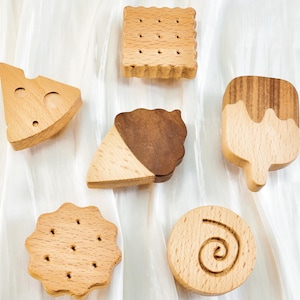 May include: A collection of six wooden food-shaped toys, including a cheese wedge, a square cracker, an ice cream cone, a popsicle, a round cookie, and a spiral-shaped cookie. The toys are made of light-colored wood with some featuring darker wood accents.