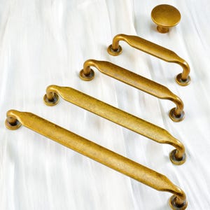 Könnte beinhalten: Eine Sammlung antiker Messing-Schrankbeschläge, darunter drei Griffleisten und ein Knauf. Die Griffe sind gebogen mit abgerundeten Enden, der Knauf ist kreisförmig. Die Beschläge haben eine warme, gealterte Oberfläche.