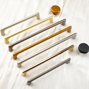 May include: A collection of cabinet hardware, including several long, straight bar pulls in various metallic finishes like gold, silver, and bronze. Also included are two round knobs, one gold and one black, all arranged on a white surface.
