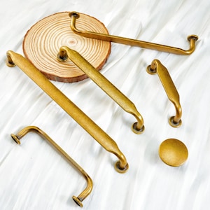 May include: A collection of antique brass cabinet hardware, including various sizes of handles and a round knob. The hardware is arranged on a white surface, with a wooden slice in the background. The hardware has a warm, golden hue.