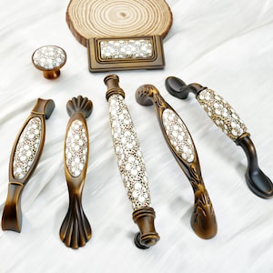 May include: A collection of decorative cabinet hardware, including knobs and pulls. The hardware features a bronze finish with white and tan floral patterned ceramic inserts. The set includes a knob, a rectangular pull, and various handle styles.