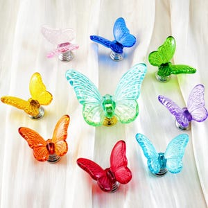 Könnte beinhalten: Eine Sammlung farbenfroher Schrankknöpfe in Schmetterlingsform. Die Knöpfe sind in verschiedenen Farben erhältlich, darunter Rosa, Blau, Grün, Gelb, Orange, Rot und Lila. Jeder Schmetterling ist auf einer silberfarbenen Basis montiert. Der Hintergrund ist ein weißer, drapierten Stoff.