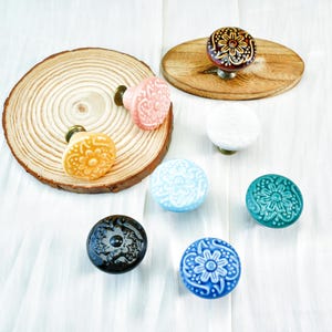 Könnte beinhalten: Eine Sammlung dekorativer Keramik-Schrankknöpfe in verschiedenen Farben und Designs. Die Knöpfe sind auf einer weißen Oberfläche angeordnet, einige davon auf Holzscheiben. Zu den Farben gehören Gelb, Rosa, Blau und Türkis.