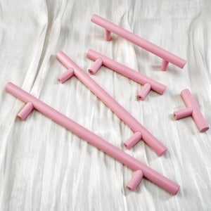 Puede incluir: Un conjunto de tiradores y manijas de armario rosas. Los herrajes son de un color rosa pastel suave. Los tiradores tienen forma de barra con extremos en forma de T. Las manijas son rectas con un extremo en forma de T a cada lado.