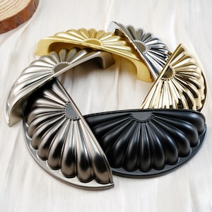 Op de afbeelding: Een verzameling decoratieve kastgrepen in diverse metallic afwerkingen, waaronder goud, zilver en zwart. Elke greep heeft een waaiervormig ontwerp met een gebogen handvat, gerangschikt in een cirkelvormig patroon op een witte ondergrond.