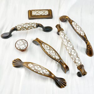 Op de afbeelding: Een verzameling decoratieve kastbeslagen, waaronder handgrepen en een knop. De hardware heeft een bronzen afwerking met witte bloemmotief keramische inzetstukken. De handgrepen variëren in vorm en grootte en bieden een reeks ontwerpopties.