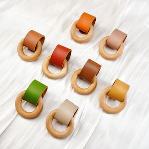 Puede incluir: Anillos de dentición de madera con correas de cuero en varios colores, incluyendo marrón, naranja, verde y beige. Los anillos están dispuestos sobre una superficie blanca texturizada.