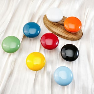 Peut inclure: Ensemble de boutons de placard brillants et colorés, verts, bleus, rouges, jaunes, noirs, blancs, oranges et bleu clair. Certains sont disposés sur un ovale en bois, le tout sur un fond de tissu blanc.