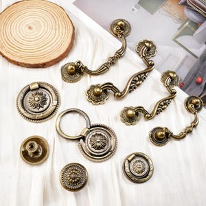 May include: An assortment of antique brass cabinet hardware, including handles, knobs, and pulls. The hardware features ornate floral designs and a warm, aged finish. A wooden slice is also present.